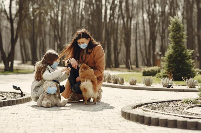happy woman with cute girl wearing protective mask and playing with adorable dog in park alley